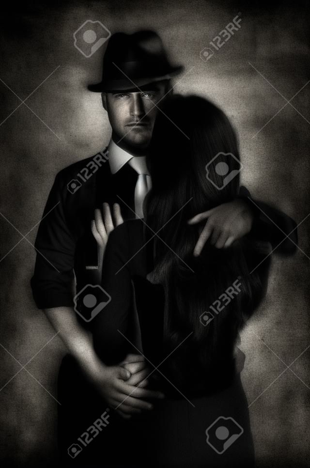 A detective with a gun and his beautiful woman leaning on him. Studio shot. Noir style. Black and white photography