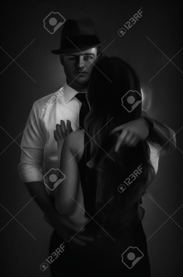 A detective with a gun and his beautiful woman leaning on him. Studio shot. Noir style. Black and white photography