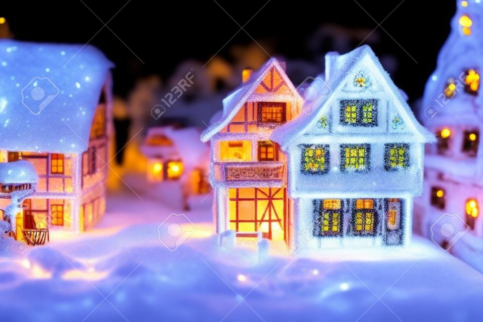 Miniaturhäuser im Dorf von Schnee bedeckt in der Nacht mit Licht Dekoration. Weihnachtswinterbaumuster für Feiertagshintergrund mit Exemplarplatz für Text.