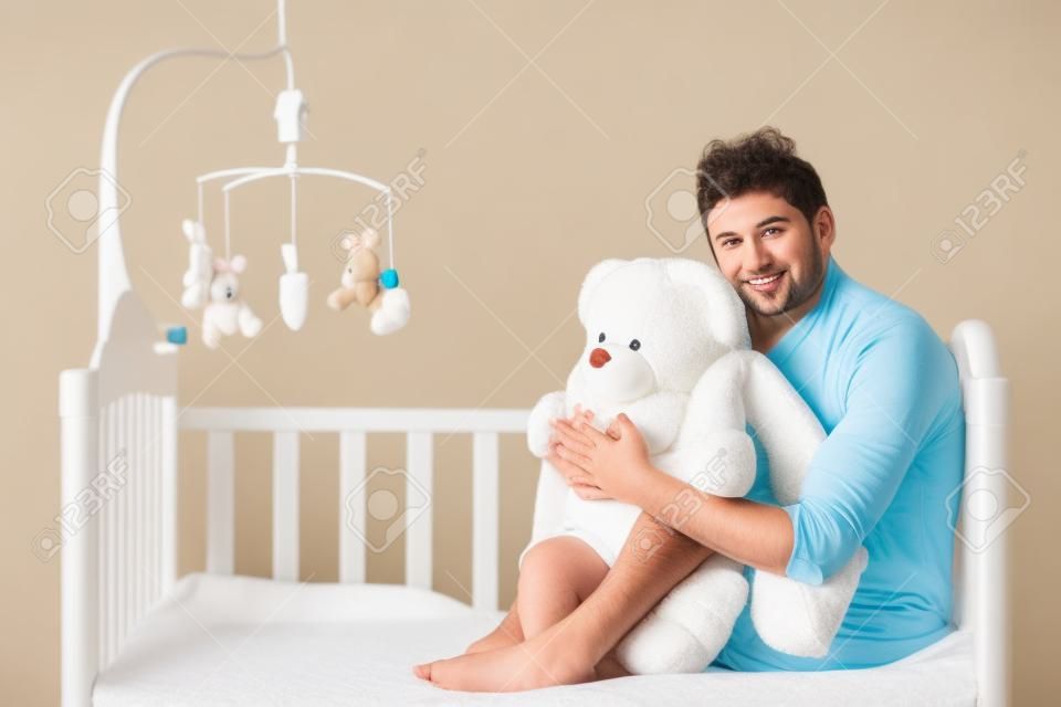 Big baby. Infant adult man in diaper holding teddy bear while sitting on the baby bed