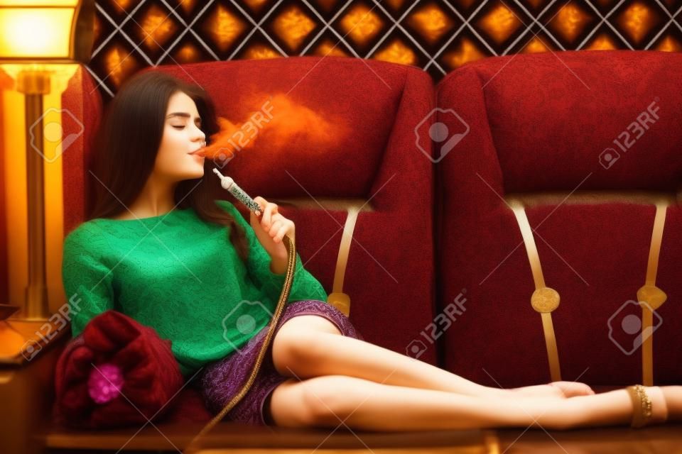 Beautiful young woman inhaling hookah. girl smoking shisha lying on sofa in cafe