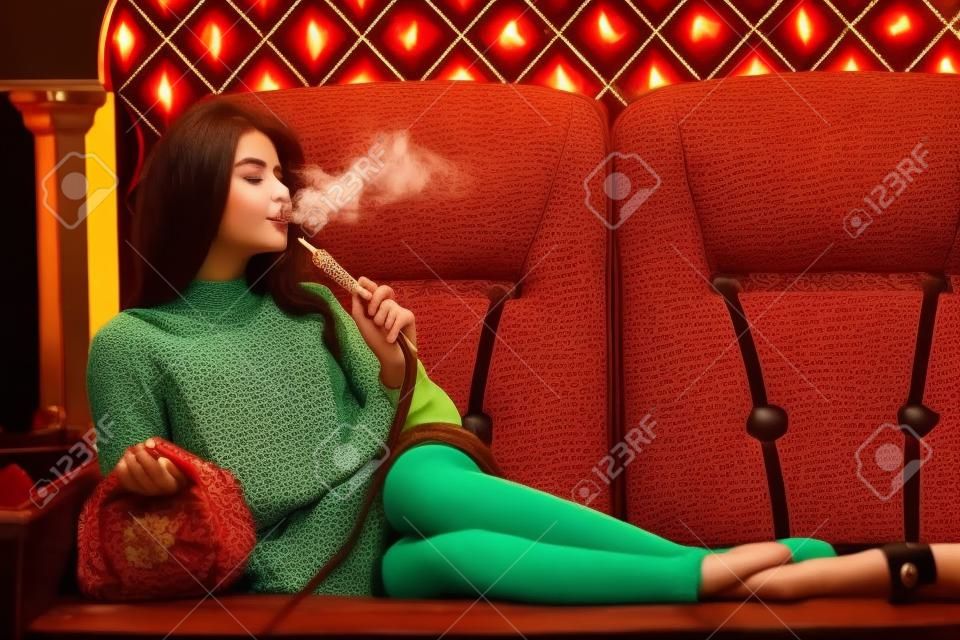 Schöne junge Frau inhalieren Wasserpfeife. Mädchen rauchen Shisha auf dem Sofa liegend im Café