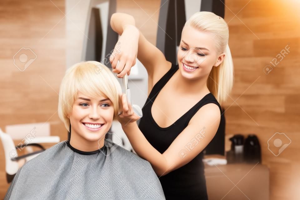 Peluquería femenino rubio cortar el cabello del hombre cliente. primer plano sobre el hombre feliz sentado en el salón de belleza y sonriente