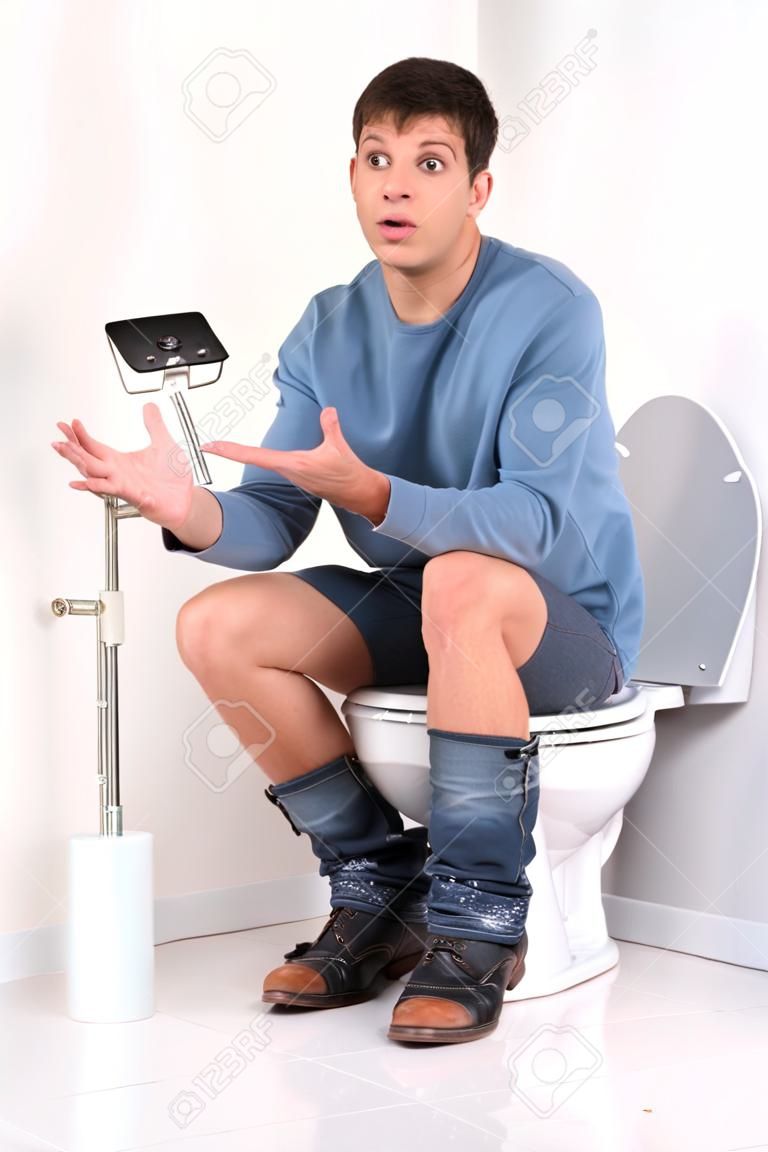 若いハンサムな男がトイレに座っています。動揺し、無力なジェスチャーをしたトイレット ペーパーなし男