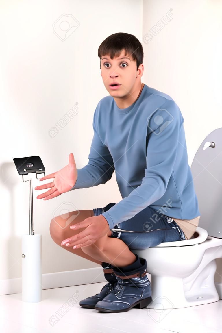 若いハンサムな男がトイレに座っています。動揺し、無力なジェスチャーをしたトイレット ペーパーなし男