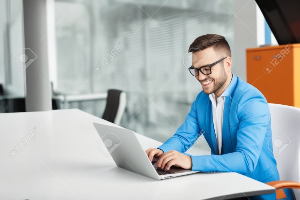 Szczęśliwy młody biznesmen za pomocą laptopa w swoim biurku.