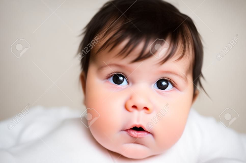 Portrait of cute sweet little newborn baby girl with black brunette hair in nice white spotted romper suit looking with big beautiful hazel brown eyes and curious glance on a bed at home. The depth of field is short.
