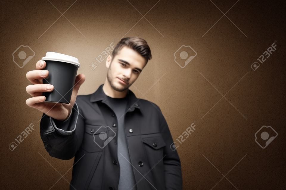 A stylish young man offers coffee. A cup of coffee in the hands of a man is sent to the camera. Cup with coffee close-up. Copyspace.