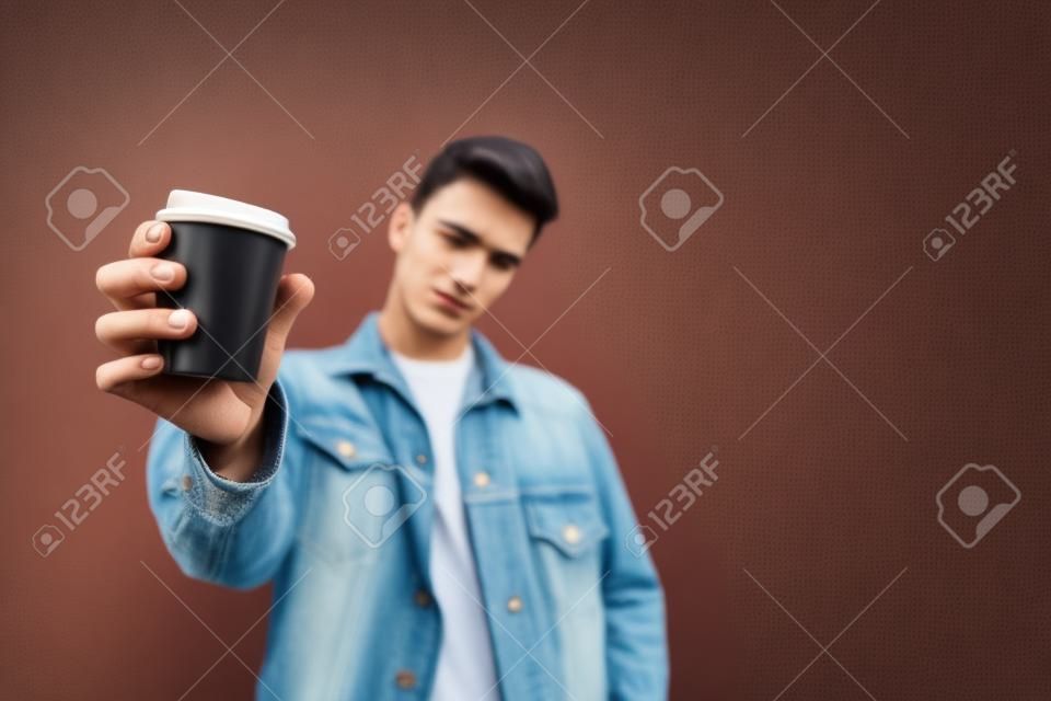 Um jovem elegante oferece café. Uma xícara de café nas mãos de um homem é enviada para a câmera. Copo com café close-up. Copyspace.
