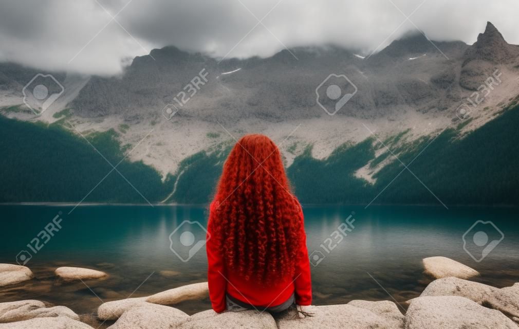 Menina de cabelos vermelhos encaracolado em uma camisa vermelha senta-se nas rochas contra o pano de fundo de um belo lago de montanha e olha para a paisagem. Turista fêmea senta-se na margem do Lago Morskie Oko, Montanhas Tatra