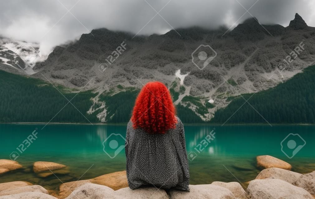 Menina de cabelos vermelhos encaracolado em uma camisa vermelha senta-se nas rochas contra o pano de fundo de um belo lago de montanha e olha para a paisagem. Turista fêmea senta-se na margem do Lago Morskie Oko, Montanhas Tatra