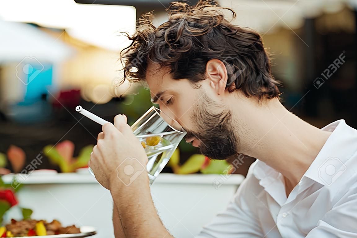 przystojny elegancki mężczyzna siedzący na zewnątrz przy stoliku w restauracji, pracujący i korzystający z laptopa podczas palenia papierosów