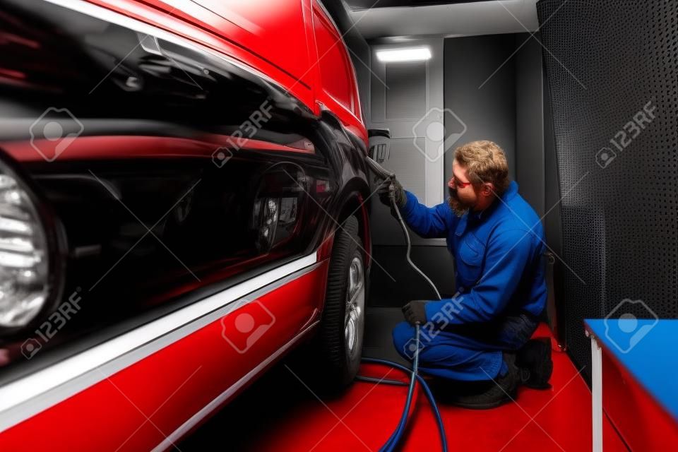 Meccanico automobilistico che lavora nell'industria manifatturiera automobilistica e che dipinge un furgone rosso in una cabina speciale