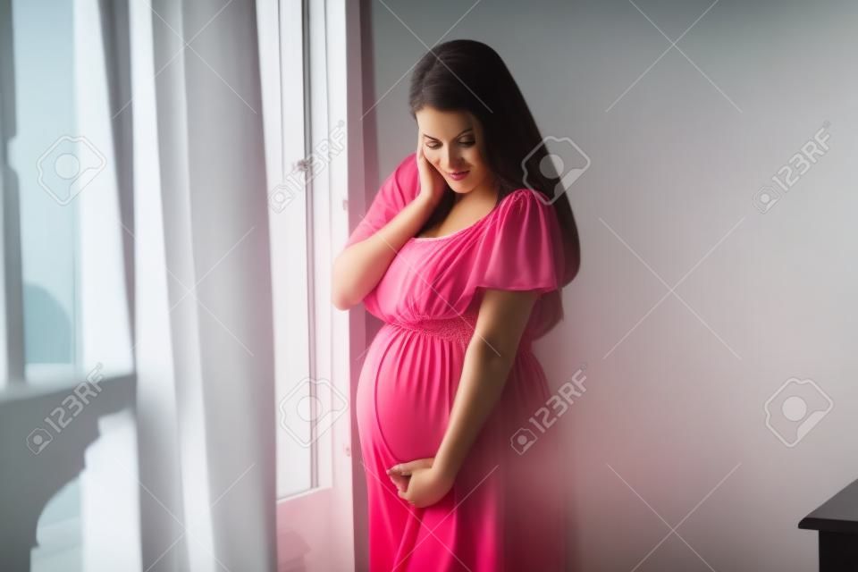 Mooie jonge zwangere vrouw die bij het raam staat