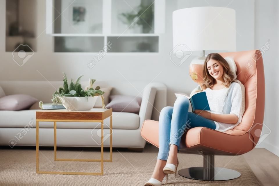 Młoda kobieta w domu siedzi na nowoczesnym krześle w pokoju i czyta książkę