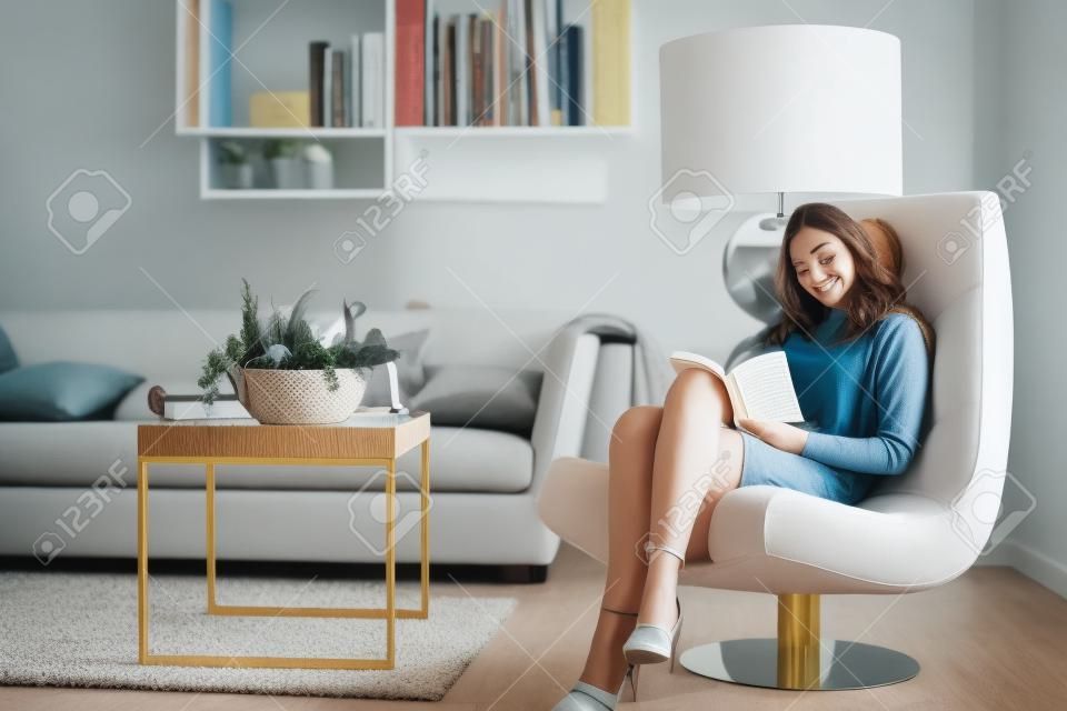 Młoda kobieta w domu siedzi na nowoczesnym krześle w pokoju i czyta książkę