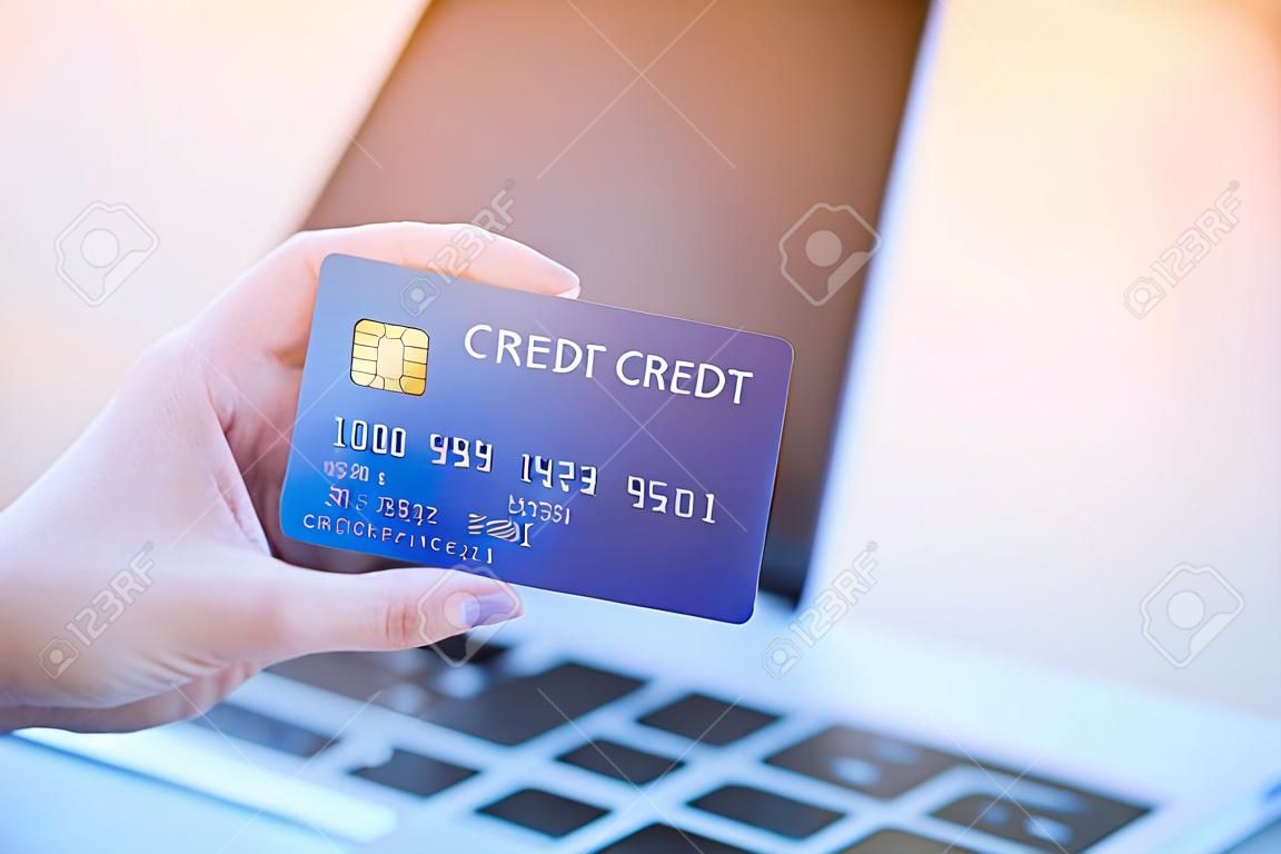 Close-up photo of left hand woman holding blue credit card mockup with a laptop background and copyspace on the right side for text. Easy e-commerce shopping online and payment by credit card