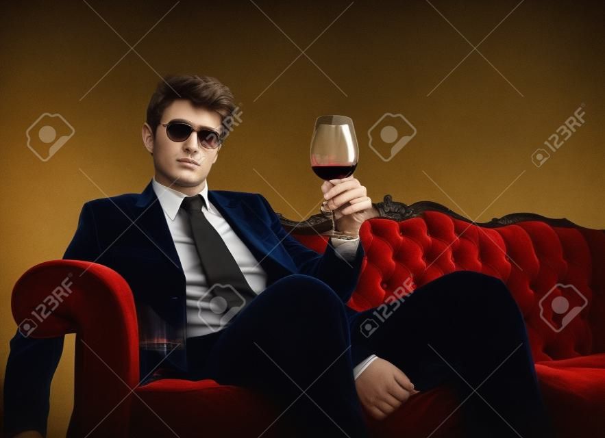 Elegante hombre sentado en un sofá de terciopelo y sosteniendo un vaso de vino