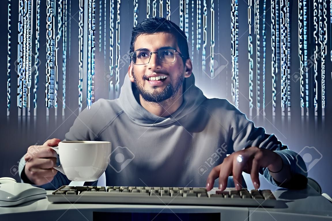 Sonriente a joven sentado frente a una pantalla de ordenador y la celebración de una taza de café