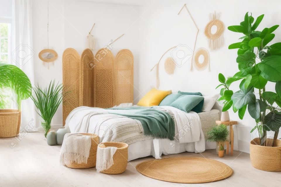 Wygodny apartament w stylu cyganerii z sypialnią hygge, poduszką i narzutą na łóżku, bambusowym parawanem, wystrojem domu, suchymi roślinami w wazonie, wiklinowym koszem, rośliną doniczkową na podłodze