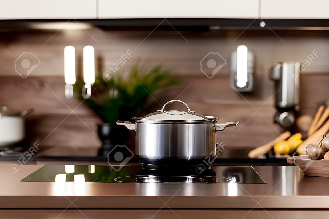 Selective focus on cooking pot on electric stove near wooden countertop against blurred background with white kitchen cupboards in modern interior. Cookery and homemade food concept