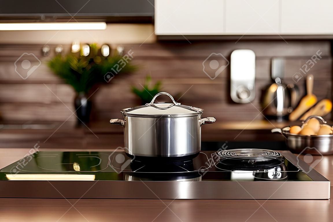 Selective focus on cooking pot on electric stove near wooden countertop against blurred background with white kitchen cupboards in modern interior. Cookery and homemade food concept
