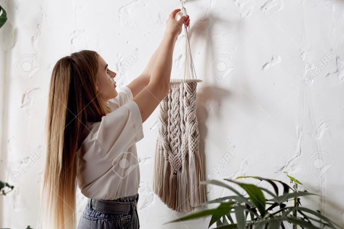 Concept of hobby and diy decoration in cozy interior. Young woman in casual clothes hangs cotton macrame on white copy space wall. Handmade knitting with natural material in boho style bedroom