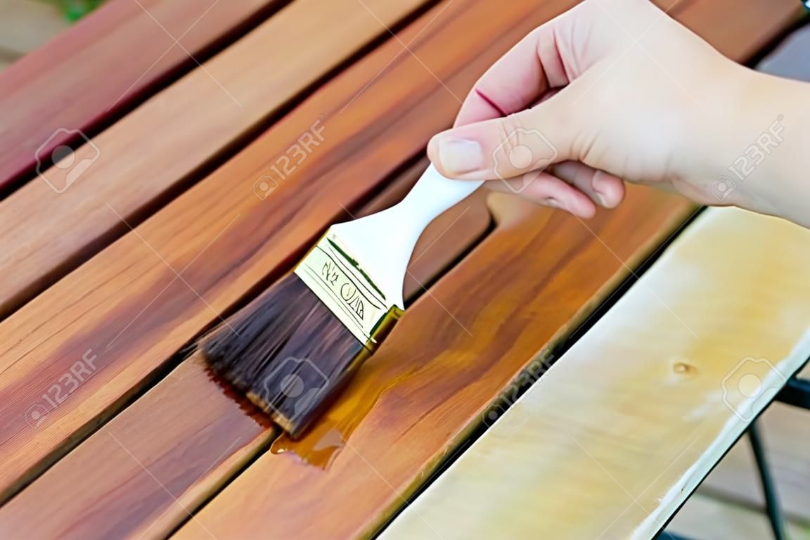 hand holding a brush applying varnish paint on a wooden garden table - painting and caring for wood with oil