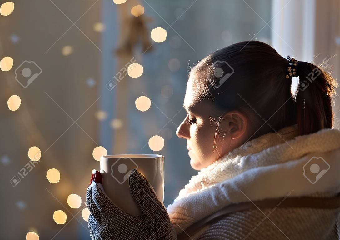 Beautiful girl covered with blanket holding cup of hot coffee with christmas lights in background, Festive moments concept
