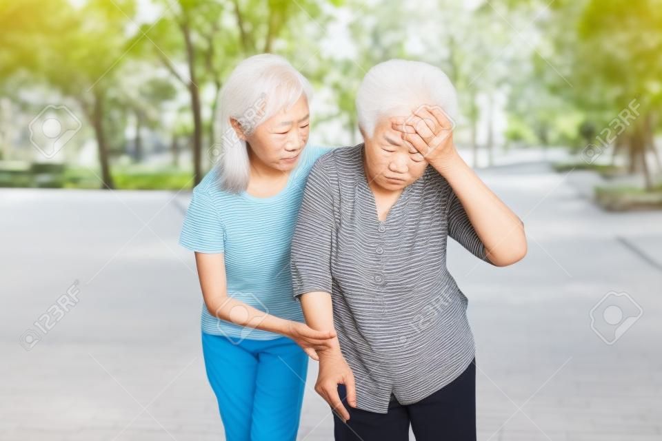 La abuela mayor asiática tiene dolor de cabeza, tocarse la cabeza con las manos, vértigo; mareos; personas mayores enfermas con presión arterial alta, se sienten desmayados, niña o nieta cuida, ayuda, apoyo en el exterior