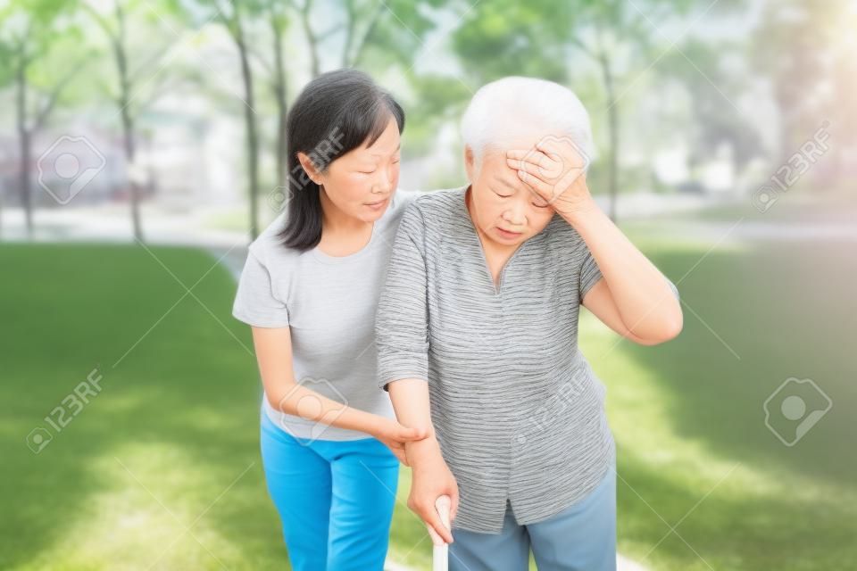 Asiatische ältere Großmutter hat Kopfschmerzen, berührt ihren Kopf mit den Händen, Schwindel, Schwindel, kranke ältere Menschen, hoher Blutdruck, Ohnmacht, Kindermädchen oder Enkelin Pflege, Hilfe, Unterstützung im Freien