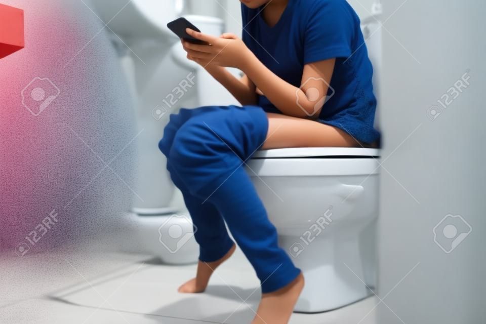 便秘のあるアジアの子供女の子、トイレでオンラインゲームをしている女性、またはソーシャルメディアの中毒、長い間便器に座っている人は痔核、ライフスタイルを引き起こす可能性があります
