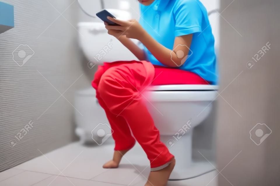 便秘のあるアジアの子供女の子、トイレでオンラインゲームをしている女性、またはソーシャルメディアの中毒、長い間便器に座っている人は痔核、ライフスタイルを引き起こす可能性があります