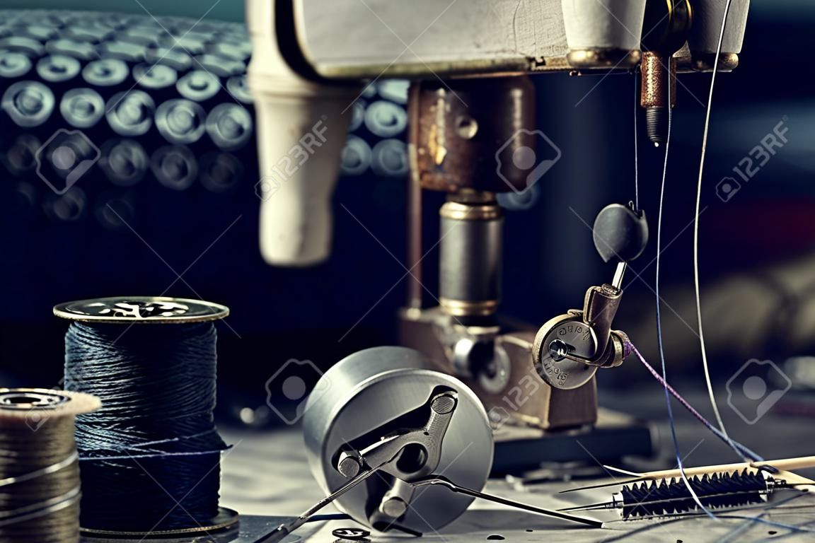 Working part of antique sewing machine with spools of threads, shuttle, measuring tape, sewing needles, scissors, a stack of fabrics on background.