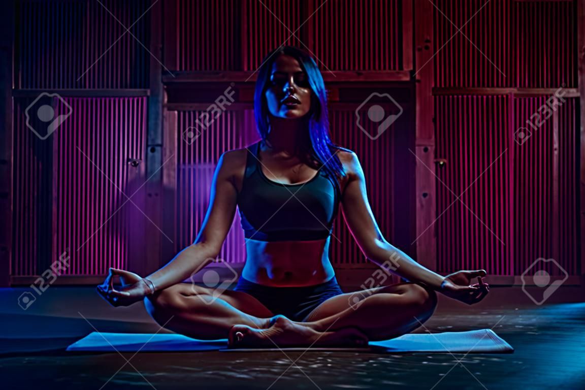 Young sport woman sitting in yoga meditation pose. Dark blue and pink night light.