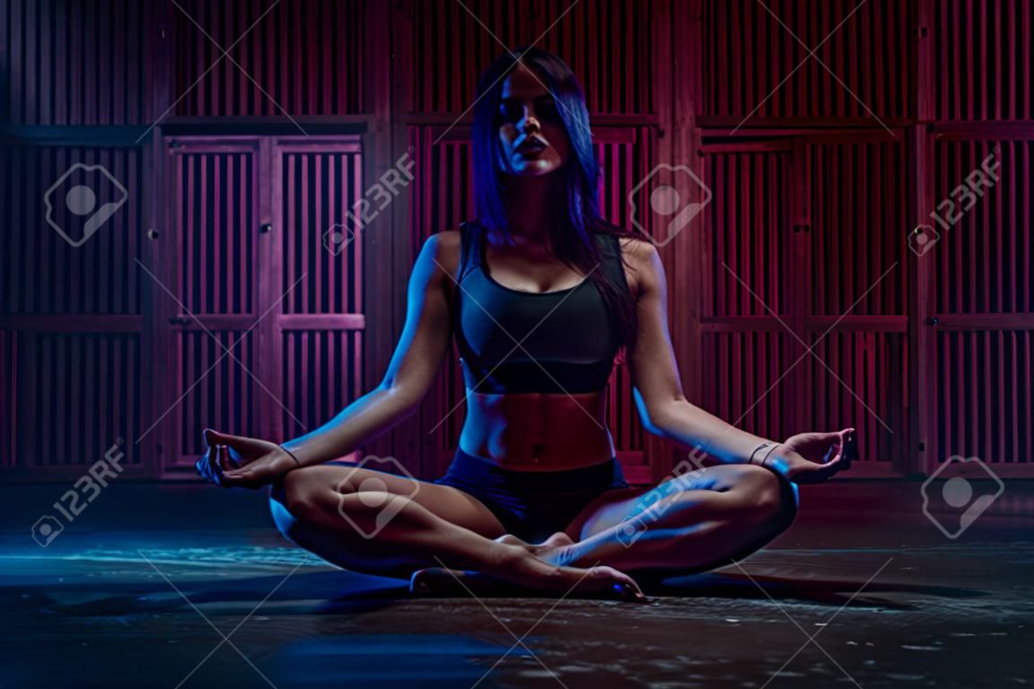 Young sport woman sitting in yoga meditation pose. Dark blue and pink night light.