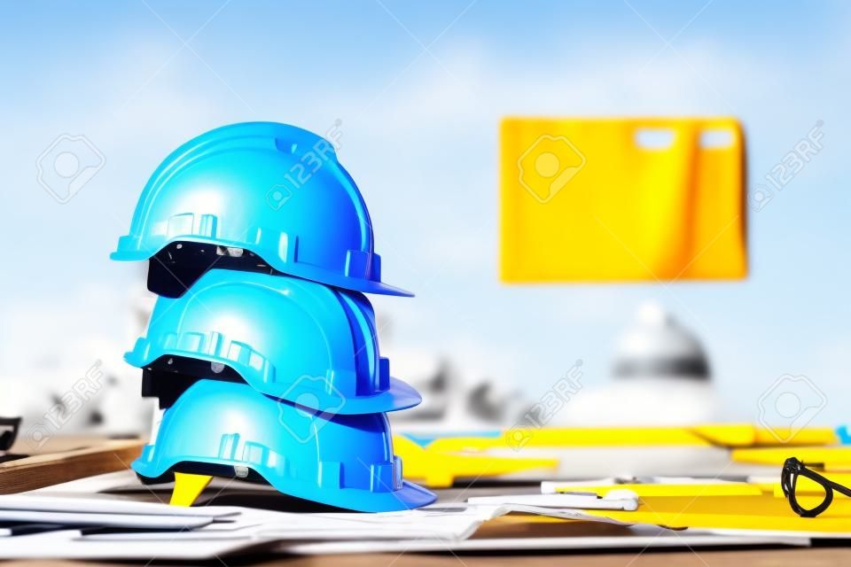 堆積在與圖紙和測量工具的桌上的白色，藍色和黃色安全帽在工程師，工頭和工作者的建造場所。安全第一的概念。