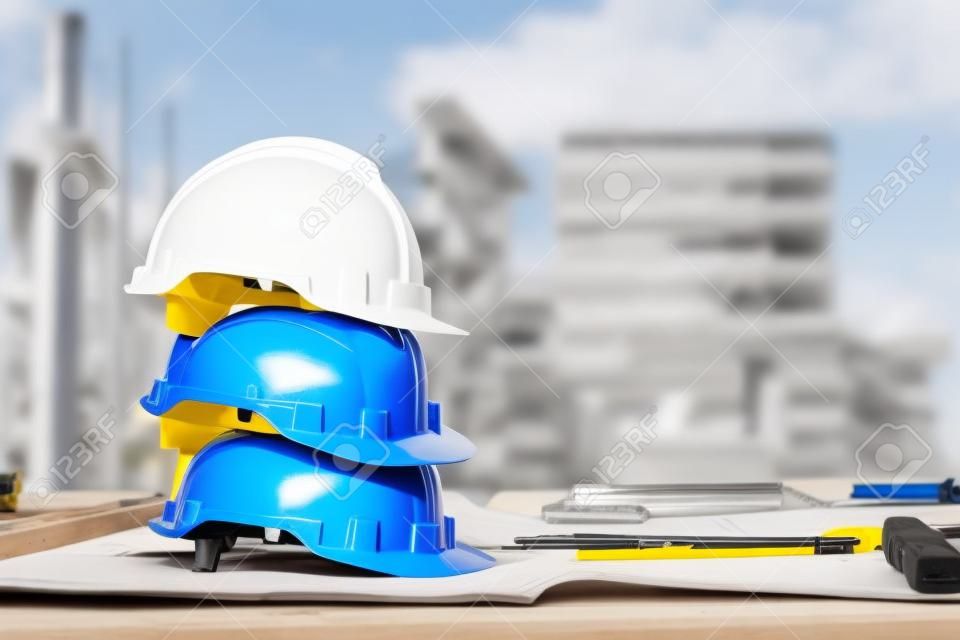 堆積在與圖紙和測量工具的桌上的白色，藍色和黃色安全帽在工程師，工頭和工作者的建造場所。安全第一的概念。