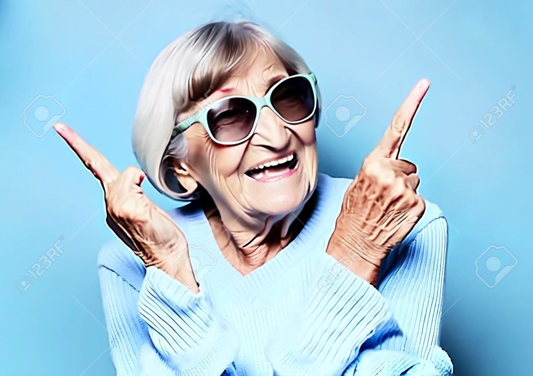 La donna anziana ride e mostra la macchina fotografica del segno della vittoria o della pace. Emozione e sentimenti. Ritratto di nonna espressiva.