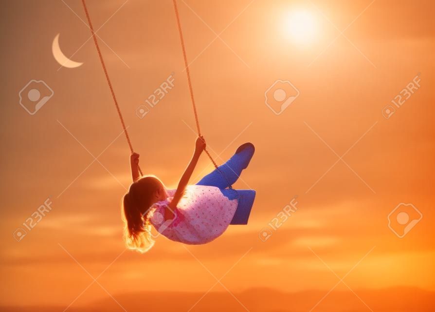 Happy child girl on swing in sunset summer