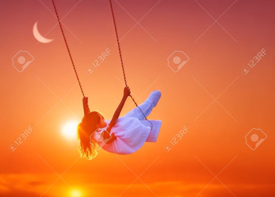 Happy child girl on swing in sunset summer
