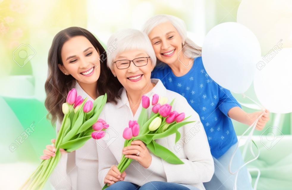 Happy mother's day! Child daughter is congratulating mom and granny giving them flowers tulips. Grandma, mum and girl smiling and hugging. Family holiday and togetherness.