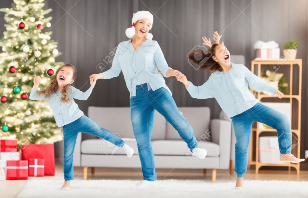 Merry Christmas, Happy Holidays. Cheerful mom and her cute daughters girls are dancing. Parent and little children having fun near tree indoors.