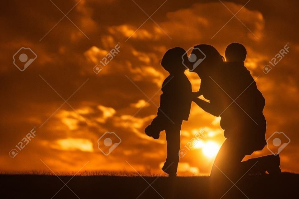 la silueta de una madre y sus dos hijos de corta edad; un niño pequeño y su hermano bebé están jugando afuera en la puesta del sol, besos y abrazos.
