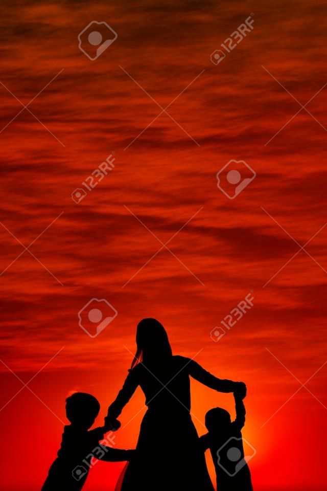 Silueta de una madre y sus dos hijos pequeños tomados de la mano y bailando alrededor de fuera, aislado contra la puesta de sol.