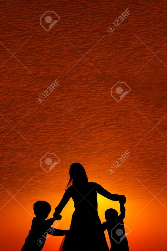Silhouette of a mother and her two young children holding hands and dancing around outside, isolated against the sunset.