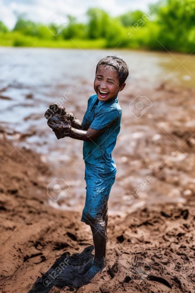 A happy young boy child is covered in mud as he laughs, swims, and plays outside in the river on a summer day.