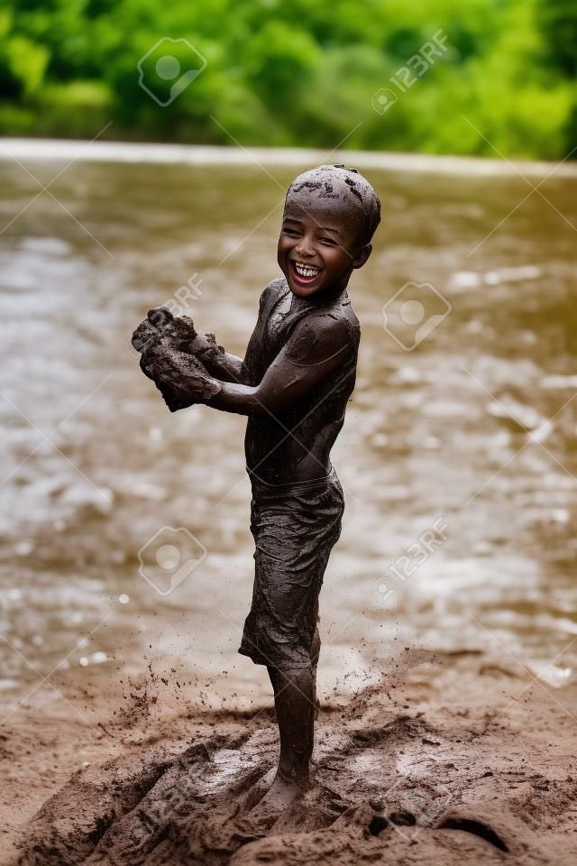 A happy young boy child is covered in mud as he laughs, swims, and plays outside in the river on a summer day.