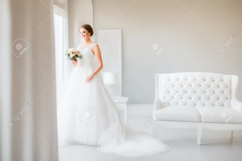 Junge Braut in einem schönen Kleid, das einen Blumenstrauß hält, der nahe Fenster in hellem weißem Studio aufwirft. Hochzeitskonzept.