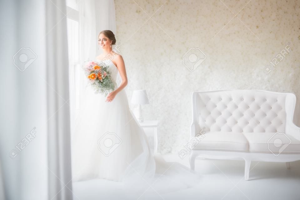 Junge Braut in einem schönen Kleid, das einen Blumenstrauß hält, der nahe Fenster in hellem weißem Studio aufwirft. Hochzeitskonzept.